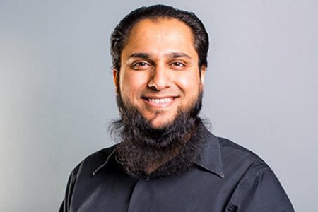 Man with a beard smiling, wearing a black shirt against a gray background.