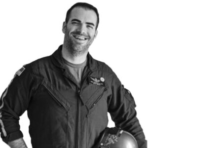 A man in a flight suit holds a helmet under his arm, smiling.