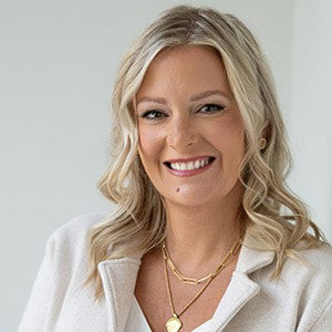 Smiling woman with wavy blonde hair wearing a white top and gold necklaces.