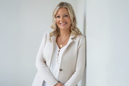 Person with wavy blonde hair wearing a light blazer and white blouse, smiling and leaning against a wall in a bright room.