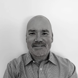 A man with a beard and a shaved head is wearing a checked shirt and smiling in a black and white photo.