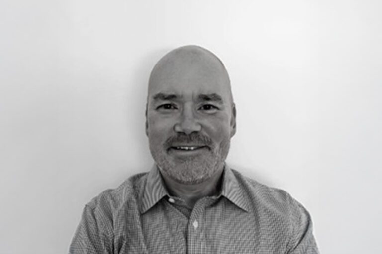 A bald man with a goatee smiles slightly, wearing a checkered shirt against a plain white background.