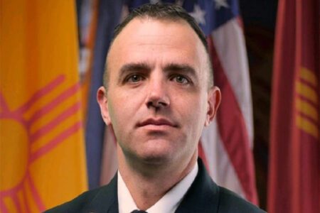 A person in a suit stands in front of several flags.