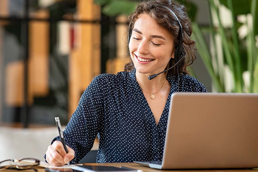 A person with a headset on, using a laptop and taking notes