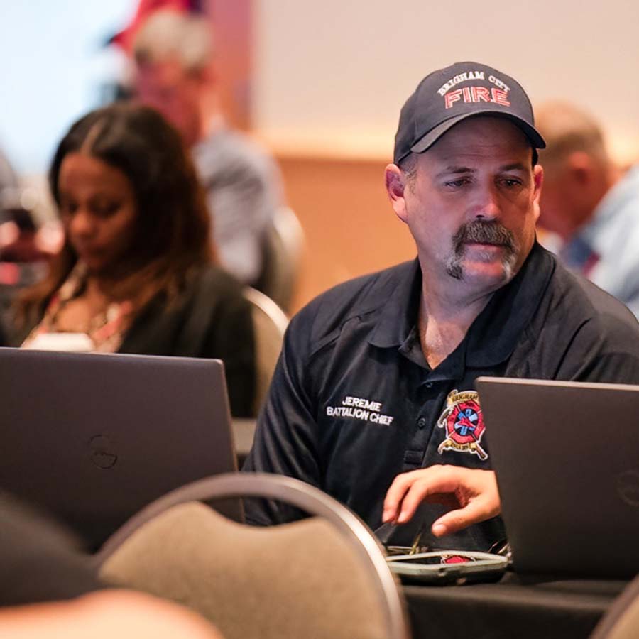 A man wearing a fire department uniform and cap sits at a table with a laptop, next to a woman. Other people are in the background.