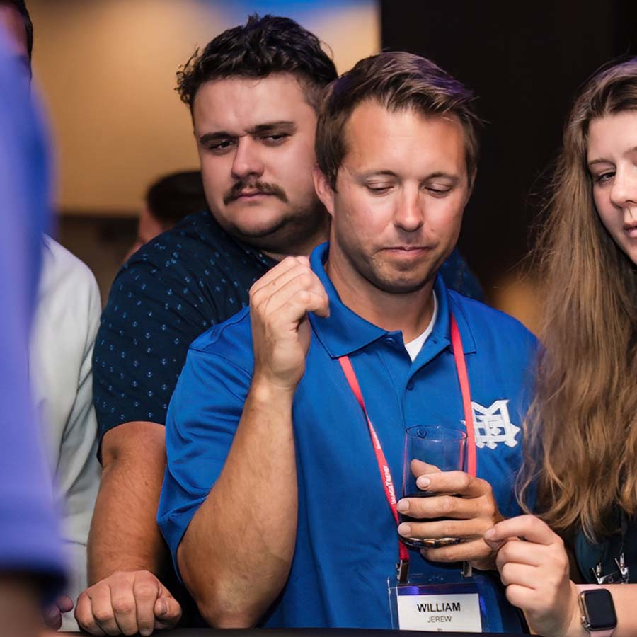 Three people are gathered at an event. A man in a blue shirt holds a drink, while another man and a woman look on. They all wear name tags.