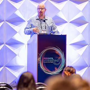 Person speaking at a podium with "Insights Unleashed" branding on the front, in front of a geometric background. Audience visible in the foreground.