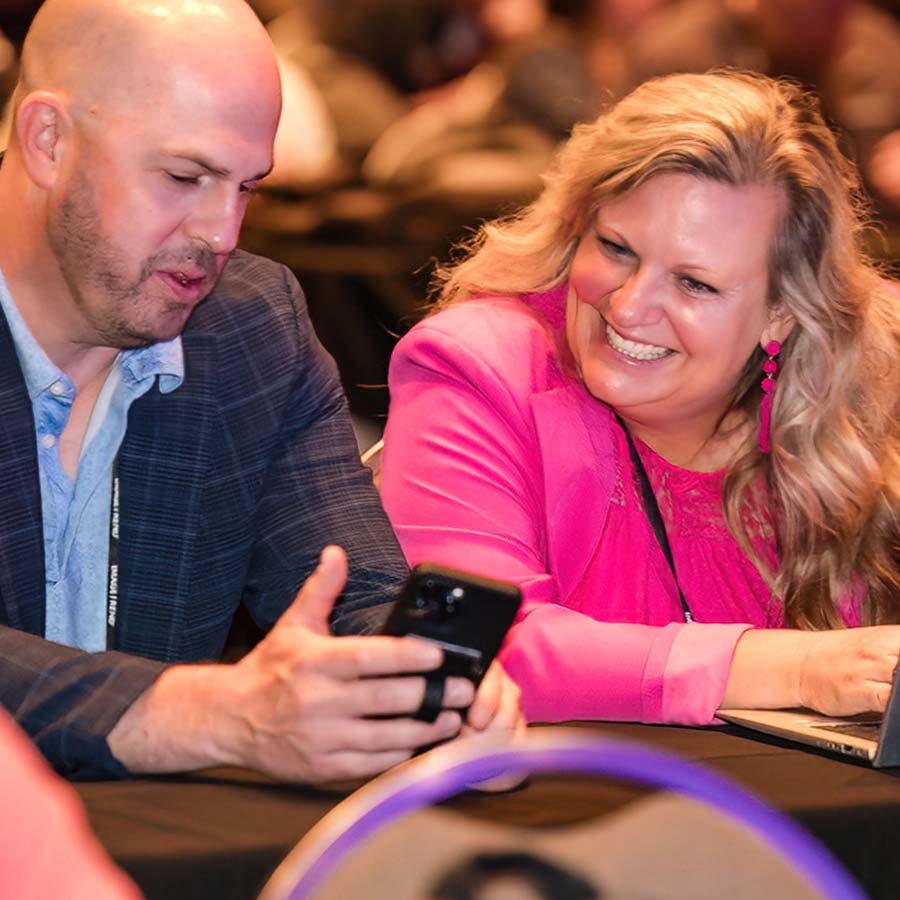 Two people sitting at a table, looking at a phone. The man wears a dark jacket, and the woman is in a pink jacket, smiling. The background is blurred, suggesting a conference setting.